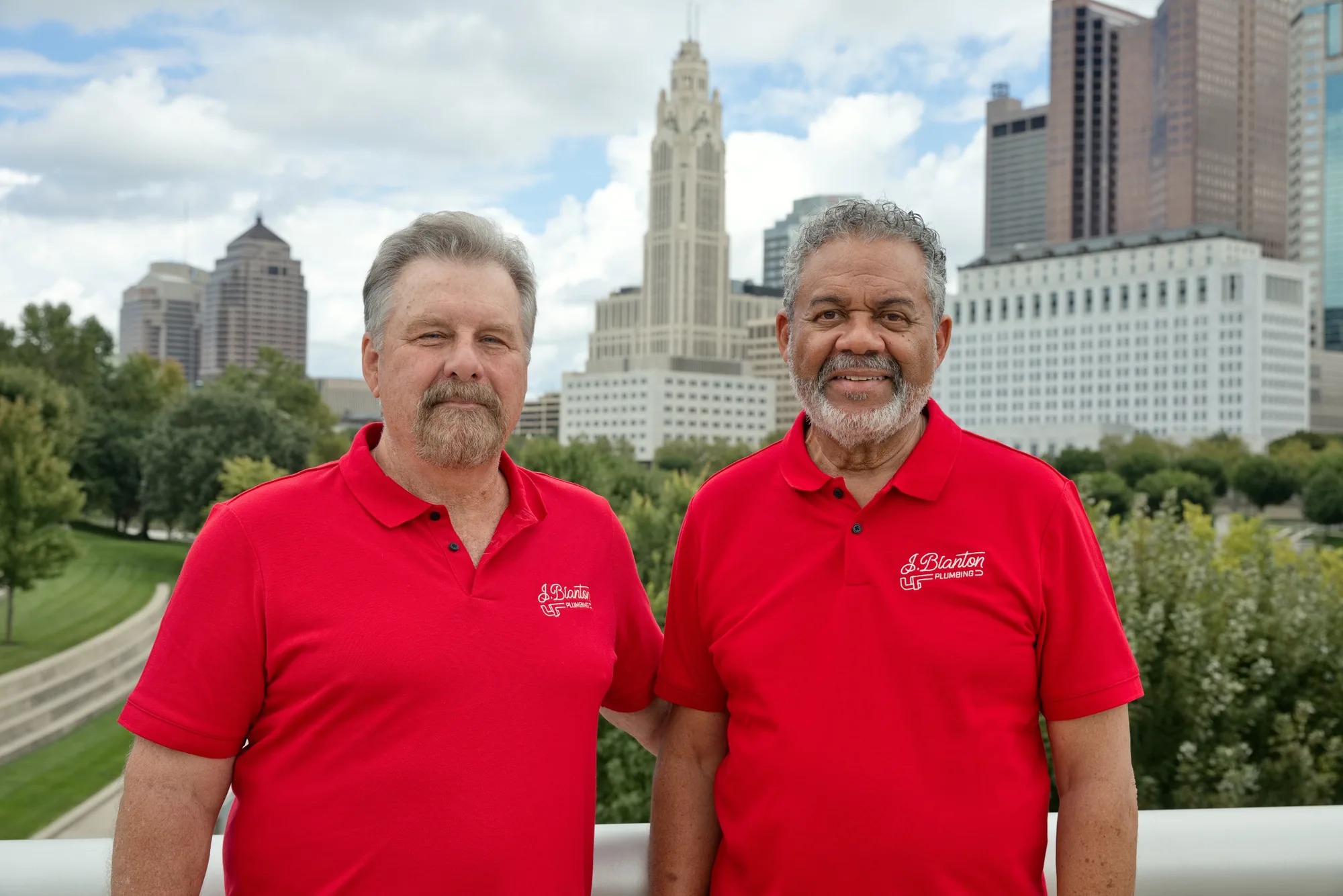 Two J. Blanton Plumbing team members in red company polos in front of the downtown Columbus skyline