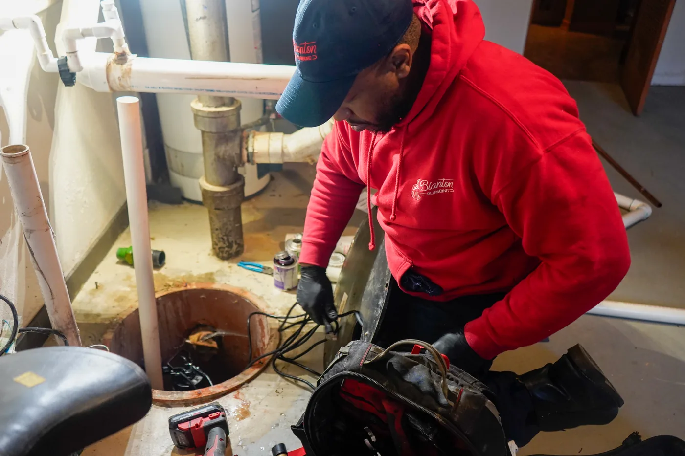 A J. Blanton Plumbing technician in a red hooded sweatshirt and work gloves kneels over an open sump pit in a basement, holding the pump’s power cord, with PVC drain lines, a water heater and a tool bag around him.